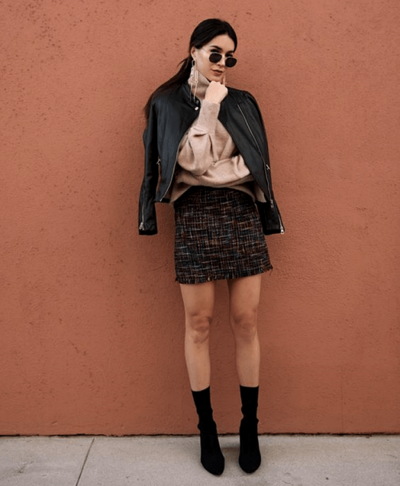 mujer con cabello oscuro usando lentes de sol, suéter beige de manga larga, chaqueta de cuero, minifalda negra tejida con líneas blancas, botines estilo sock boots negras