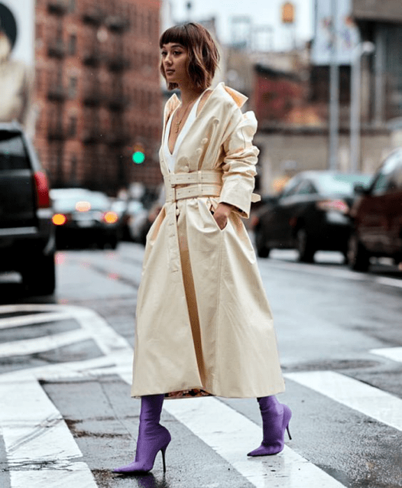 mujer con cabello castaño corto usando un top de tirantes blanco, abrigo largo color beige, botas estilo sock boots moradas de satén