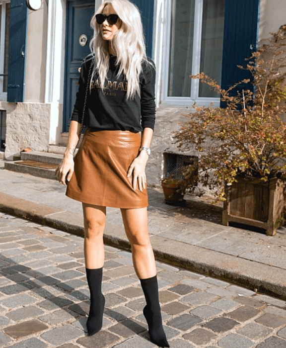 mujer rubia platinada usando lentes de sol, sudadera negra balmain, minifalda de cuero café, botines estilo sock boots negros