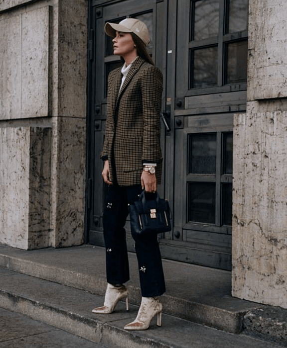 mujer con cabello castaño usando una gorra beige, top blanco de cuello alto, blazer café de cuadros negros, pantalón negro de vestir con estrellas, botines beige de terciopelo estilo sock boot y bolso de mano negro