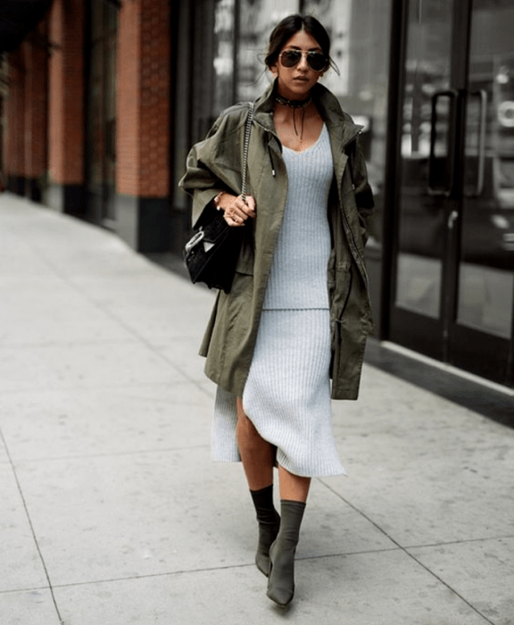 mujer morena usando lentes de sol, vestido tejido gris, chaqueta larga color verde militar, bolso negro de mano y botines estilo sock boots verde militar