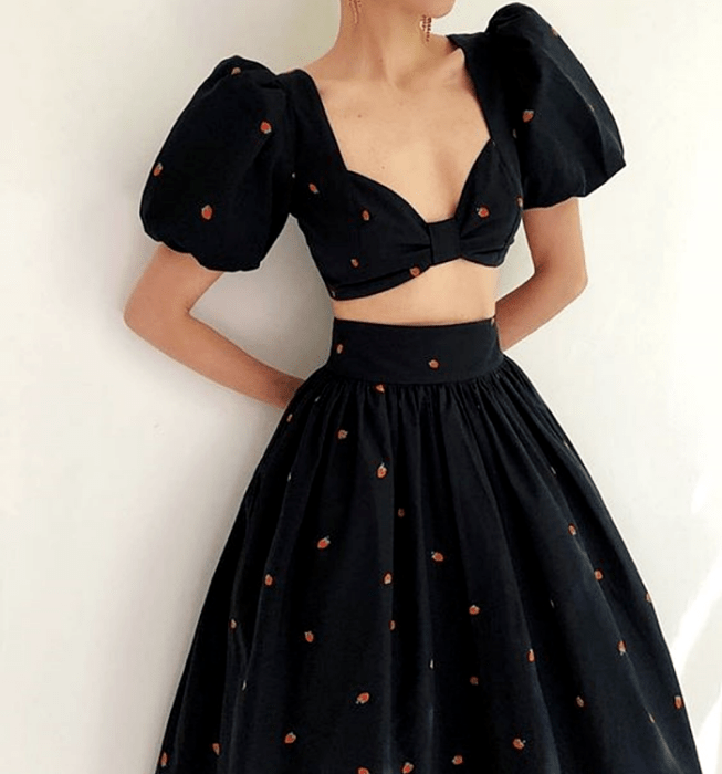 chica con un vestido negro de dos piezas, top de mangas cortas abombadas y escote profundo, falda larga con volumen, estampada con fresas pequeñas