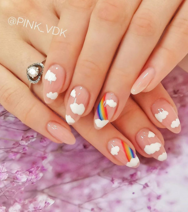 Manicura con diseño de cielo y nubes; uñas largas en forma de almendra pintadas con esmalte nude y transparente con una figura de arcoíris