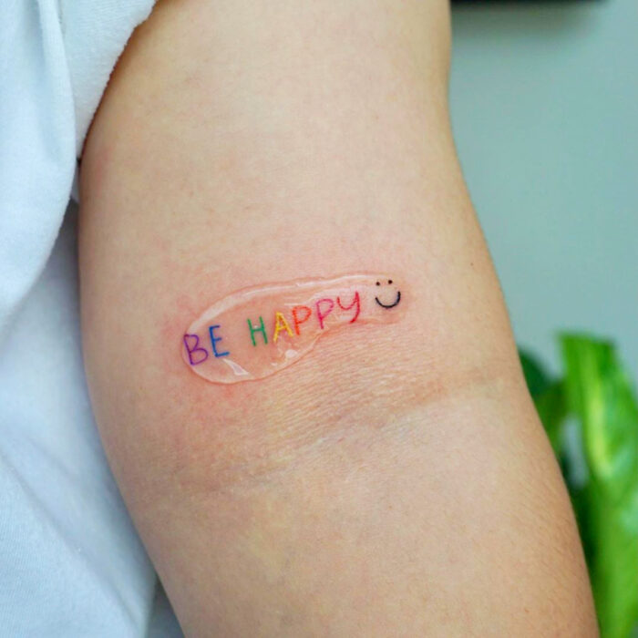 Diseños motivadores de tatuajes con las palabras Be Happy de colores arcoíris en el brazo