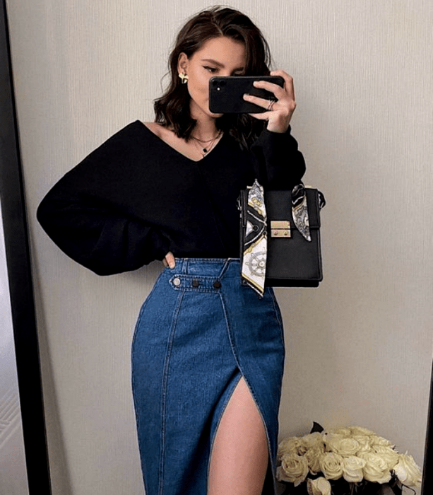 chica de cabello negro usando una blusa negra de mangas abombadas, falda de mezclilla larga con abertura en la pierna y bolso de mano negro con blanco