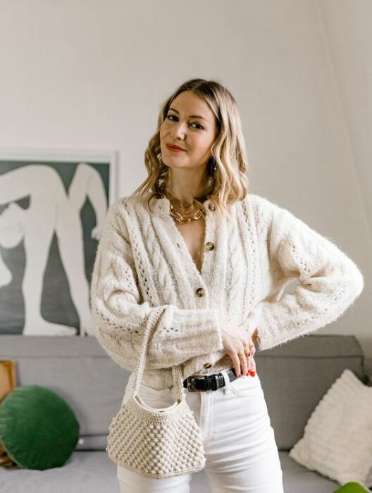 Chica rubia de melena corta con rizos, pantalón blanco, suéter de punto con botones blanco y bolso tejido claro junto a accesorios dorados
