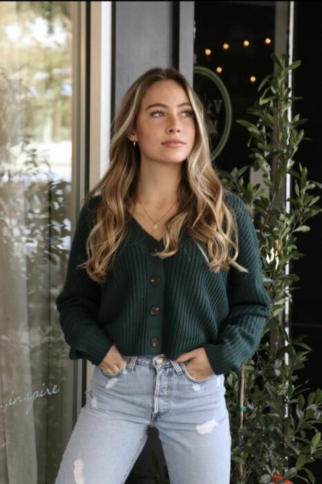 Chica rubia de cabello largo y ondas suaves, vistiendo jeans claros y un suéter verde con botones
