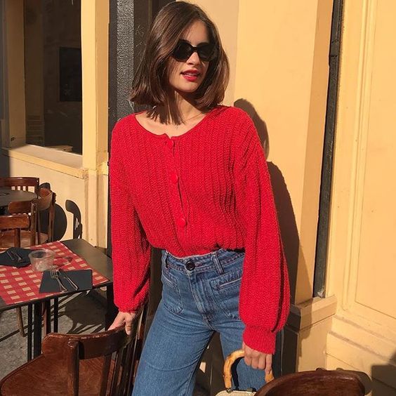 Chica delgada con melena corta, lentes de sol, labial rojo, vistiendo jeans oscuros y suéter de punto rojo