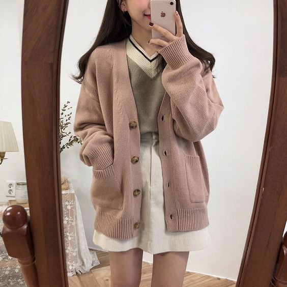 Chica asiática tomándose una selfie frente al espejo, vistiendo una falda de pana blanca, suéter gris y otro suéter rosa palo encima con botones carey