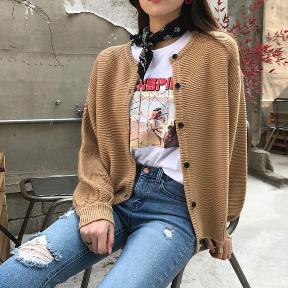Chica sentada en una silla gris, vestida con jeans oscuros, blusa blanca, suéter beige amplio de punto con botones negros y un pañuelo en el cuello