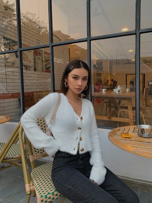 Chica delgada sentada en una mesa al exterior de una cafetería, vistiendo pantalón negro, suéter blanco con botones cafés y cabello muy corto con broches al costado