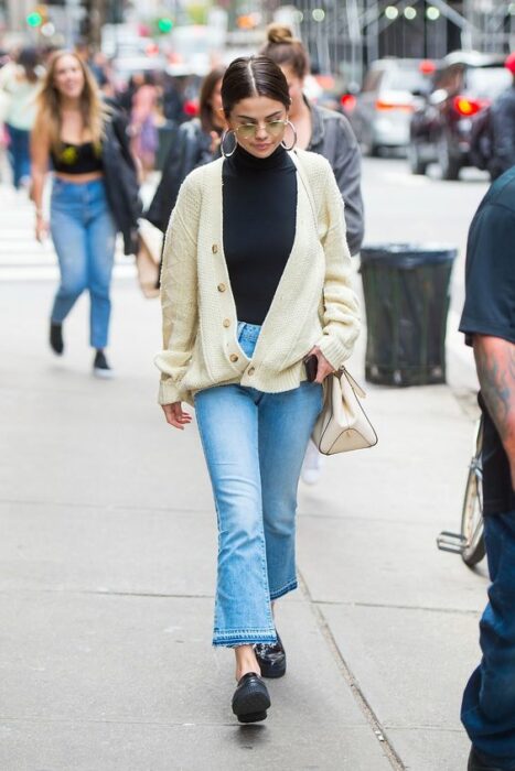 Selena Gómez caminando en la calle, vistiendo pantalón de mezclilla acampanado, zapatos negros, blusa negra de cuello y suéter largo tejido con botones beige