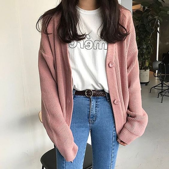 Chica asiática de cabello oscuro con jeans oscuros, blusa blanca y suéter rosa largo tejido con botones rosa