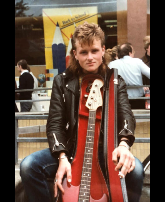 Foto vintage, fotografía antigua en color, de hombre rockero de cabello corto de arriba y largo de abajo, rubio, con bajo, chamarra de cuero negra