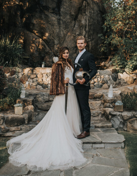 Ideas para una boda vikinga; novios, esposos en el bosque con vestido blanco y largo y traje negro, con casco con cuernos