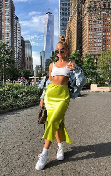 Chica rubia con chongo alto vestida con top blanco, chamarra de mezclilla, falda de satín verde limón y tenis blancos