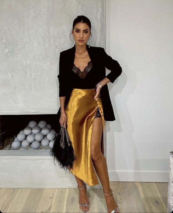 Mujer morena con coleta alta vestida con blusa de encaje, saco negro, falda amarilla de satín y tacones dorados