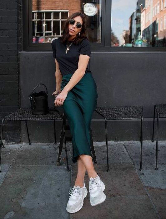 Chica de cabello largo y negro ondulado sentada en una banca, vistiendo blusa negra con falda de satín verde esmeralda y tenis blancos