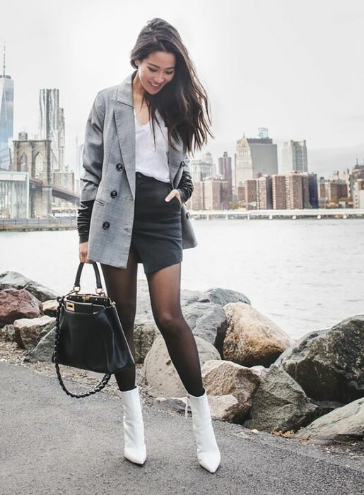 Chica usando botines blancos y medias negras, mini falda de vinil, blusa blanca además de blazer gris con bolso de mano negro