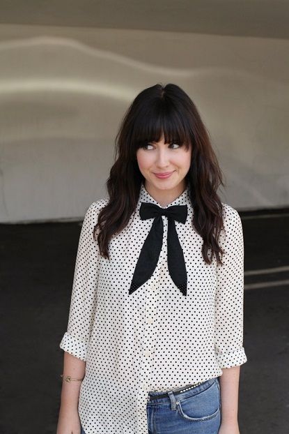 Chica con cabello largo y oscuro con ondas y flequillo, vistiendo jeans, blusa blanca con puntos negros y moño negro en el cuello