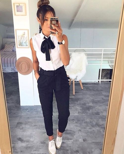 Chica tomándose un selfie frente al espejo con pantalones oscuros, blusa blanca sin mangas y moño negro en el cuello