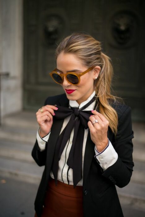 Chica rubia con coleta y gafas de sol, vistiendo una falda cuadros, camisa blanca, saco negro y un moño negro en el cuello
