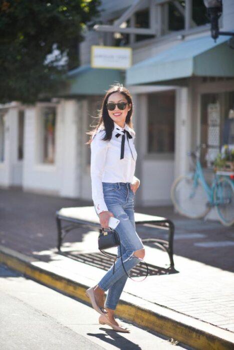 Chica delgada de cabello medio negro y lacio, vistiendo jeans rotos, camisa blanca y moño negro en el cuello
