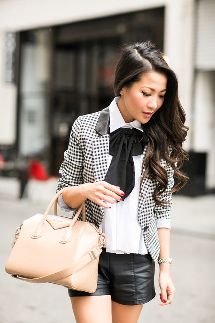 Chica asiática de cabello largo, negro y ondulado, sosteniendo una bolsa beige, vistiendo short de cuero negro, camisa blanca y moño negro en el cuello