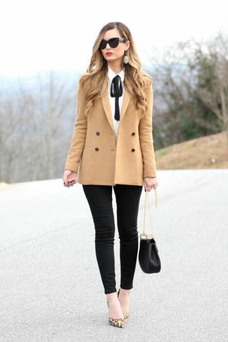 Chica rubia con cabello suelto y gafas de sol, vistiendo pantalón negro, camisa blanca, abrigo beige y moño negro en el cuello
