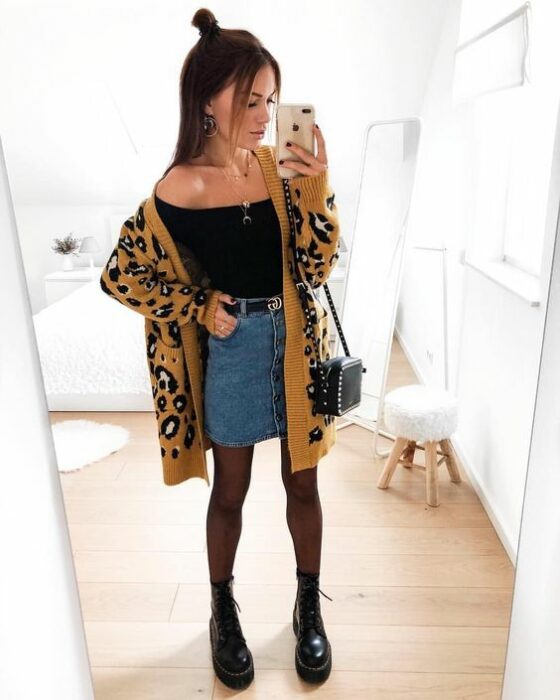 Chica usando blusa negra, mini falda denim, con medias y botines negros y un cardigan largo de estampado animal print