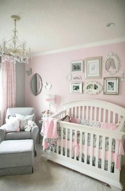 Decoración de la habitación de una bebé en tonos rosa con un sofá en la esquina de color gris y diferentes cuadros sobre la cuna color blanco