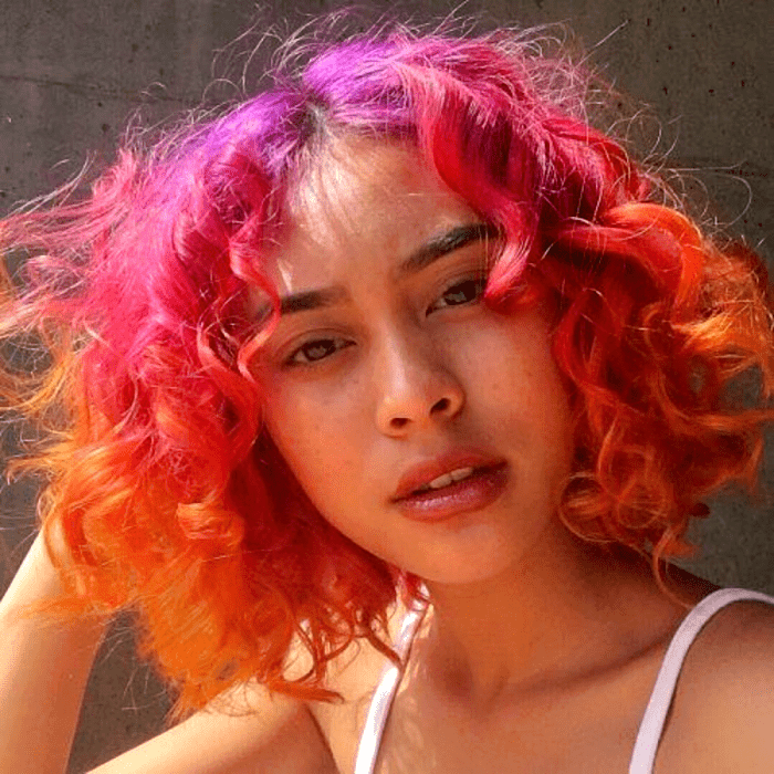 chica morena con cabello chino teñido color rosa, naranja y rojo neón estilo ombré, usando un top blanco de tirantes