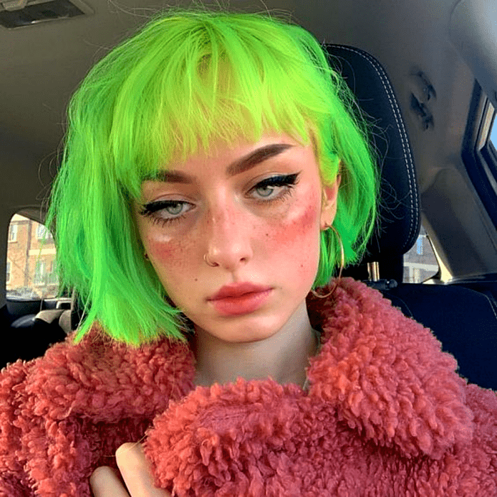 chica de cabello verde neón corto, usando maquillaje dramático con rubor exagerado y delineador negro grueso. usando un abrigo tipo teddy color rosa