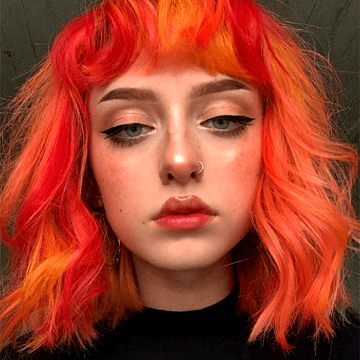 chica de cabello naranja y rojo brillante, neón con fleco corto y ondas. usando una camiseta negra de cuello redondo y delineador negro grueso