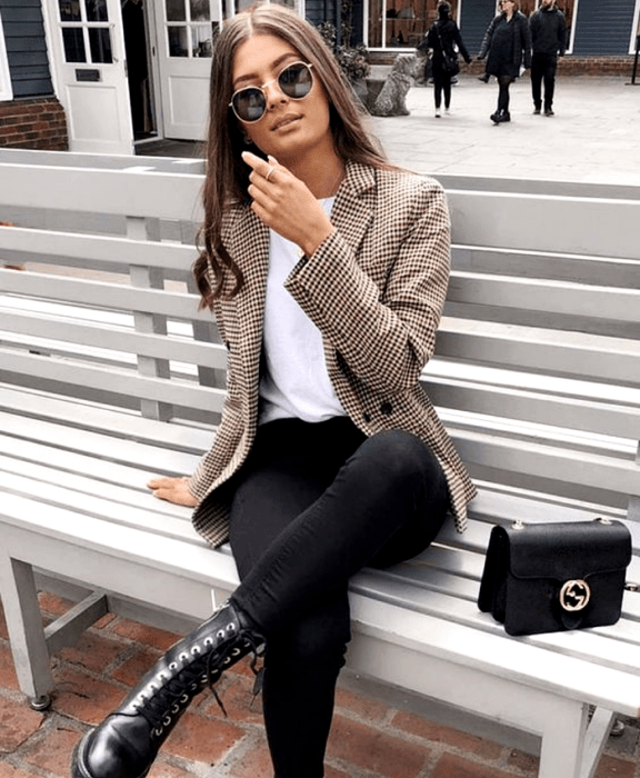 chica de cabello castaño claro usando lentes de sol, camiseta blanca, blazer café beige de cuadros, skinny jeans negros, botines negros de piel y bolso mini negro