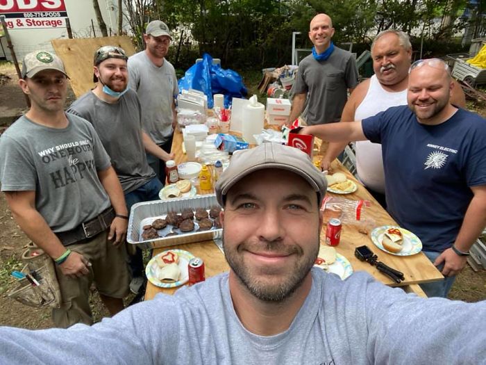 John Kinney tomando una selfie con un grupo de constructores detrás de él al rededor de una mesa rectangular de madera con comida encima