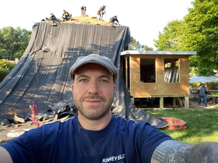 John Kinney tomando una selfie vistiendo una blusa azul marino y de fondo la reparación de una casa