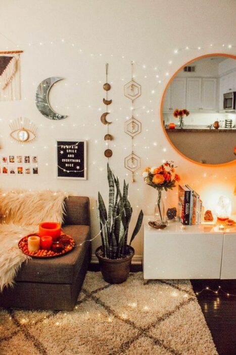 Habitación acogedora de paredes blancas con colgantes de luna, espejo redondo, mueble blanco para libros y florero, una planta verde y un sillón café con una tele de peluche blanco encima
