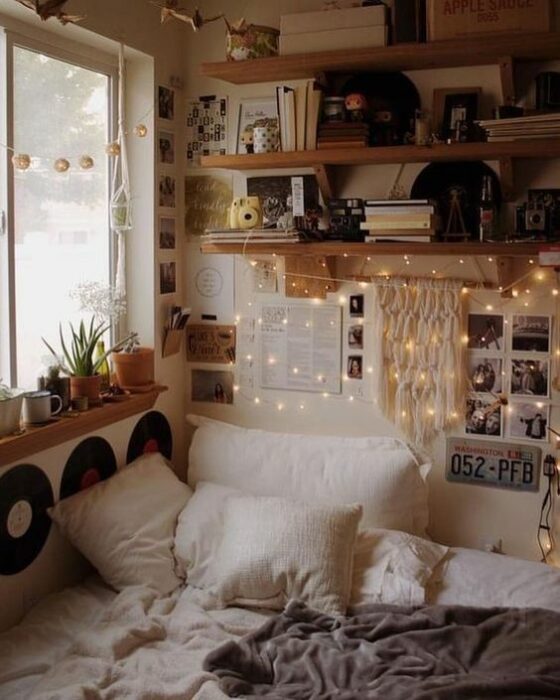Habitación acogedora con cama en una esquina pegada a la ventana con cobijas y cojines blancos y la pared blanca con repisas de madera llenas de discos y libros y series de luces amarillas colgando
