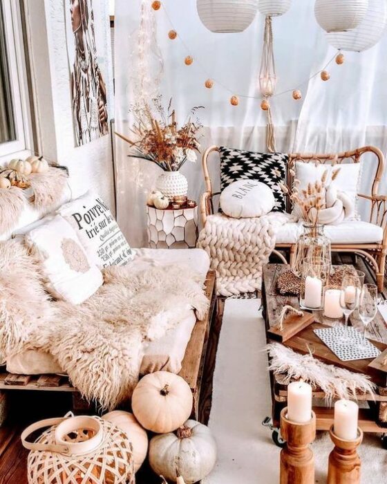 Habitación acogedora en tonos rosas con un sillón blanco cubierto por una tela de peluche blanca y decorado con dos cojines; junto a un sillón de madera con cojín blanco y una manta tejida beige; en el centro hay una mesa con velas y decoración de calabazas