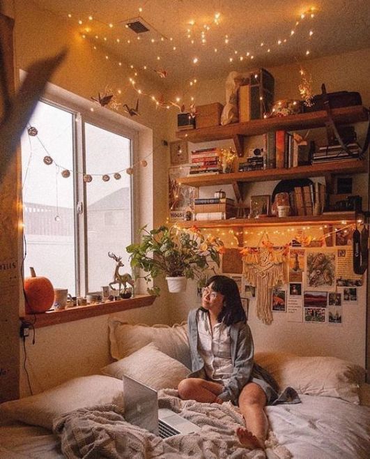 Chica asiática con cabello corto mirando hacia el cielo, sentada en una cama de cobijas blancas pegada a la ventana. A su espalda, fotos pegadas y estantes con libros y luces amarillas