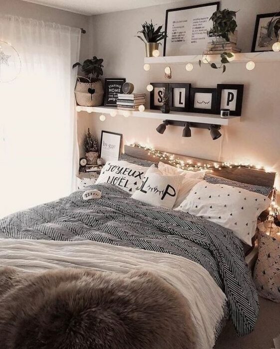 Habitación luminosa con paredes blancas y una cama en la esquina del cuarto; cobijas blancas con detalles de flores rosas; en la pared hay fotografías, cuadros de mariposas y repisas con flores, además de luces amarillas rodeando la cama