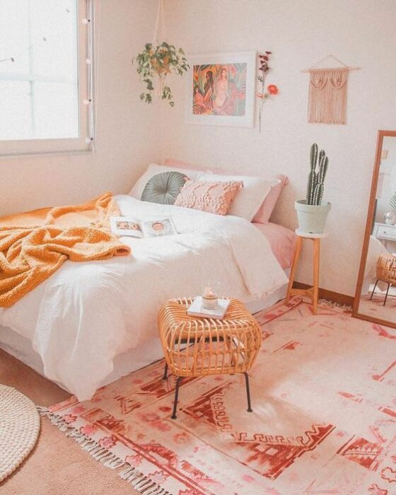 Habitación acogedora con paredes claras, cuadros y plantas colgantes, y en la esquina, una cama con colcha blanca, cojines rosas y azules, frazada naranja en la orilla, además de un espejo de cuerpo completo y una alfombra blanca con rosa en el piso