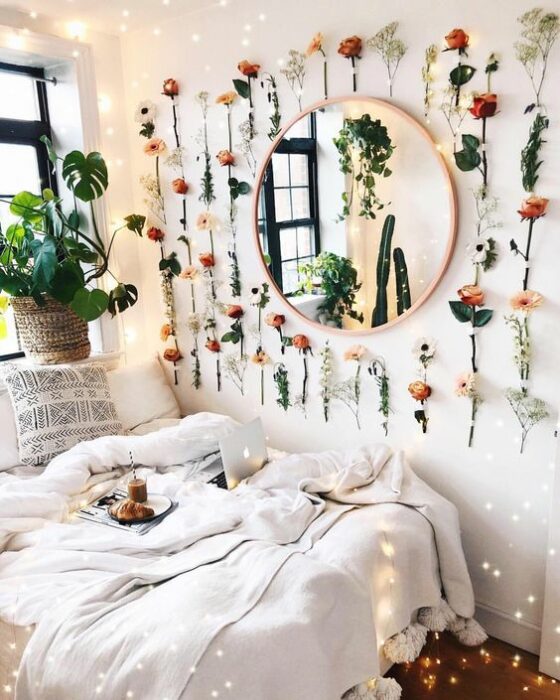 Habitación con paredes blancas y un espejo redondo rodeado de flores naturales pegadas en la pared. Abajo, una cama con cobijas blancas junto a la ventana