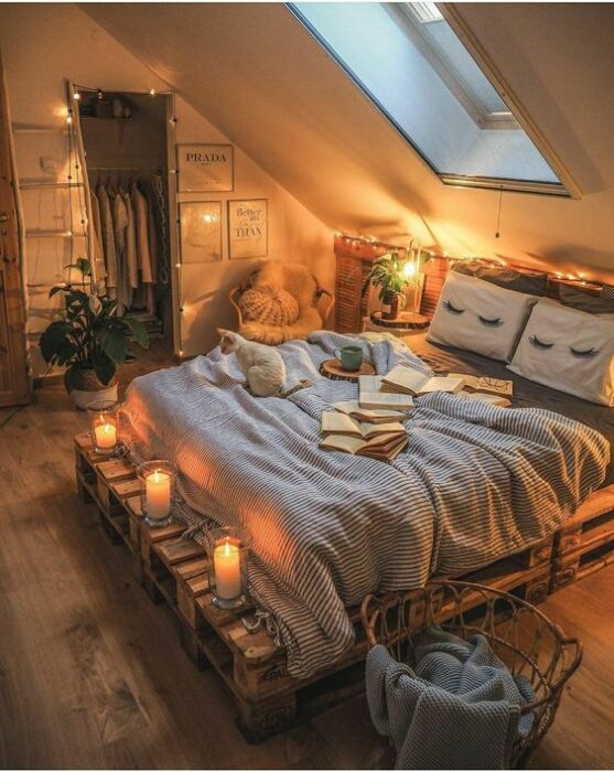 Habitación acogedora con tragaluz; la cama tiene base de pallets y cobijas grises con cojines blancos y paredes blancas