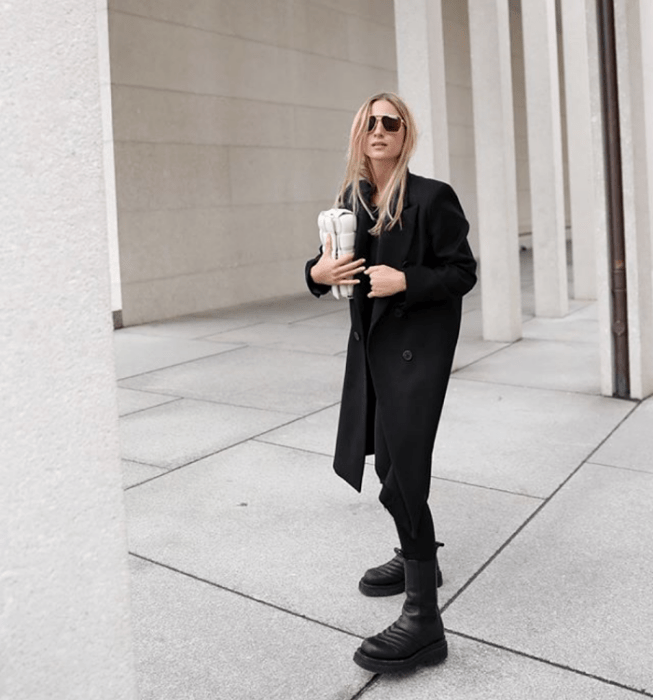 chica rubia usando lentes de sol, abrigo largo negro, top negro, leggings negros, botas de plataforma negras y bolso de mano beige