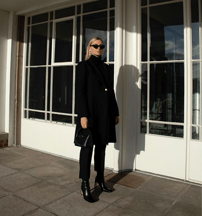 chica rubia con lentes de sol, abrigo largo negro, suéter negro de cuello alto, pantalón de vestir negro, botines negros de tacón y bolso de mano negro