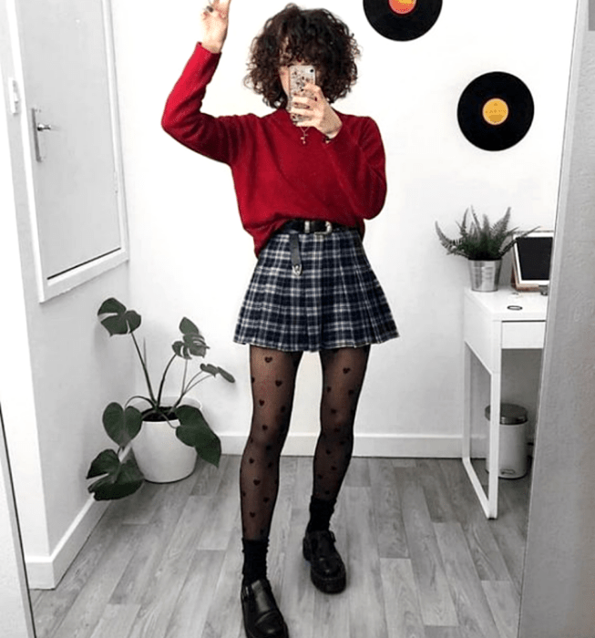 Chica de cabello chino usando un suéter rojo de manga larga, minifalda de tablones escocesa con cuadros azules y blancos, medias negras con corazones, cinturón negro y mocasines negros de suela gruesa
