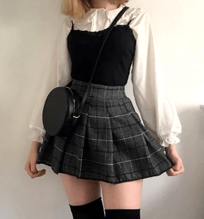 Chica rubia usando una camisa blanca de mangas abombadas, top negro de tirantes, minifalda gris de cuadros estilo escocesa con tablones, medias negras largas y bolso negro crossbody