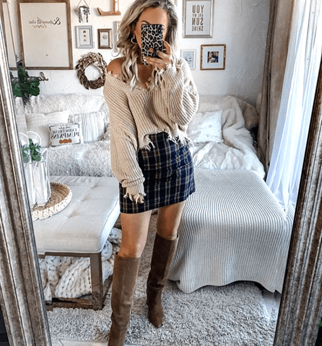 Chica rubia platinada usando un suéter beige con escote en V, minifalda azul marino de cuadros estilo escocesa, botas cafés largas con tacón